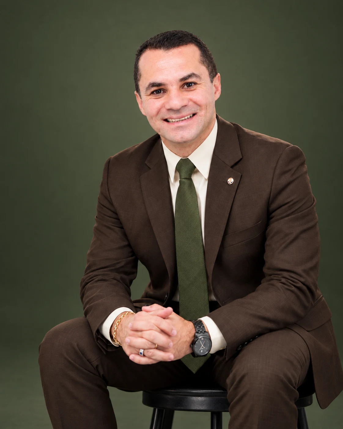 Adriano Arestides sentado, sorrindo, de terno marrom e gravata verde-musgo